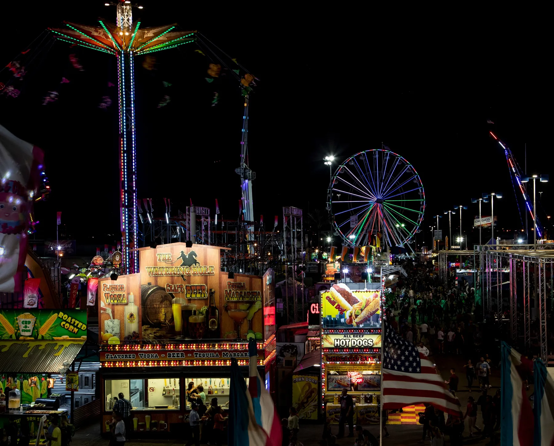 The Arizona State Fair midway at night: a lit Ferris wheel, a spinning swing ride, food stands and crowds under an American flag