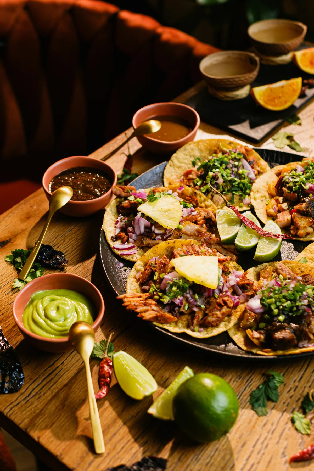Tacos al pastor topped with pineapple, onion and cilantro on a platter, with bowls of salsa and avocado salsa, lime wedges and dried chiles on a wooden restaurant table