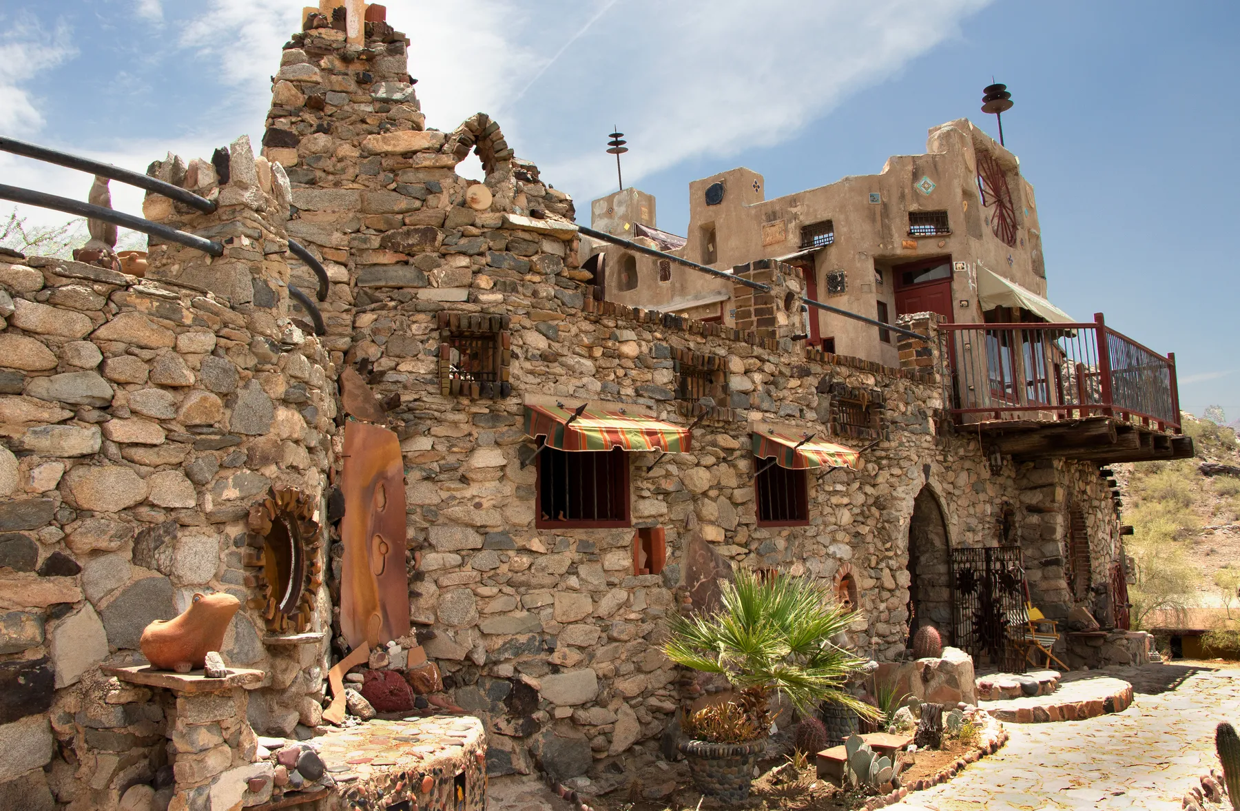 Hand-stacked stone walls and towers of Mystery Castle in Phoenix under a blue sky, with striped awnings, barred windows, a wagon wheel set into an adobe tower and a red-railed balcony