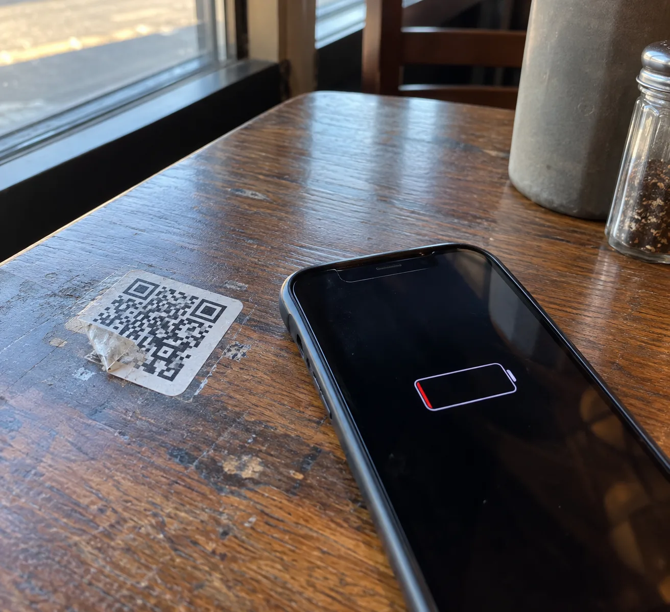 A peeling QR code sticker on a restaurant table next to a phone with a dead battery