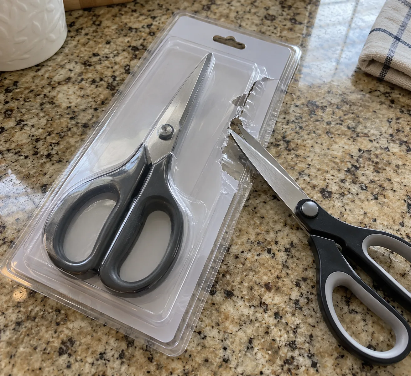 New scissors sealed in a plastic clamshell, next to a second pair cutting it open